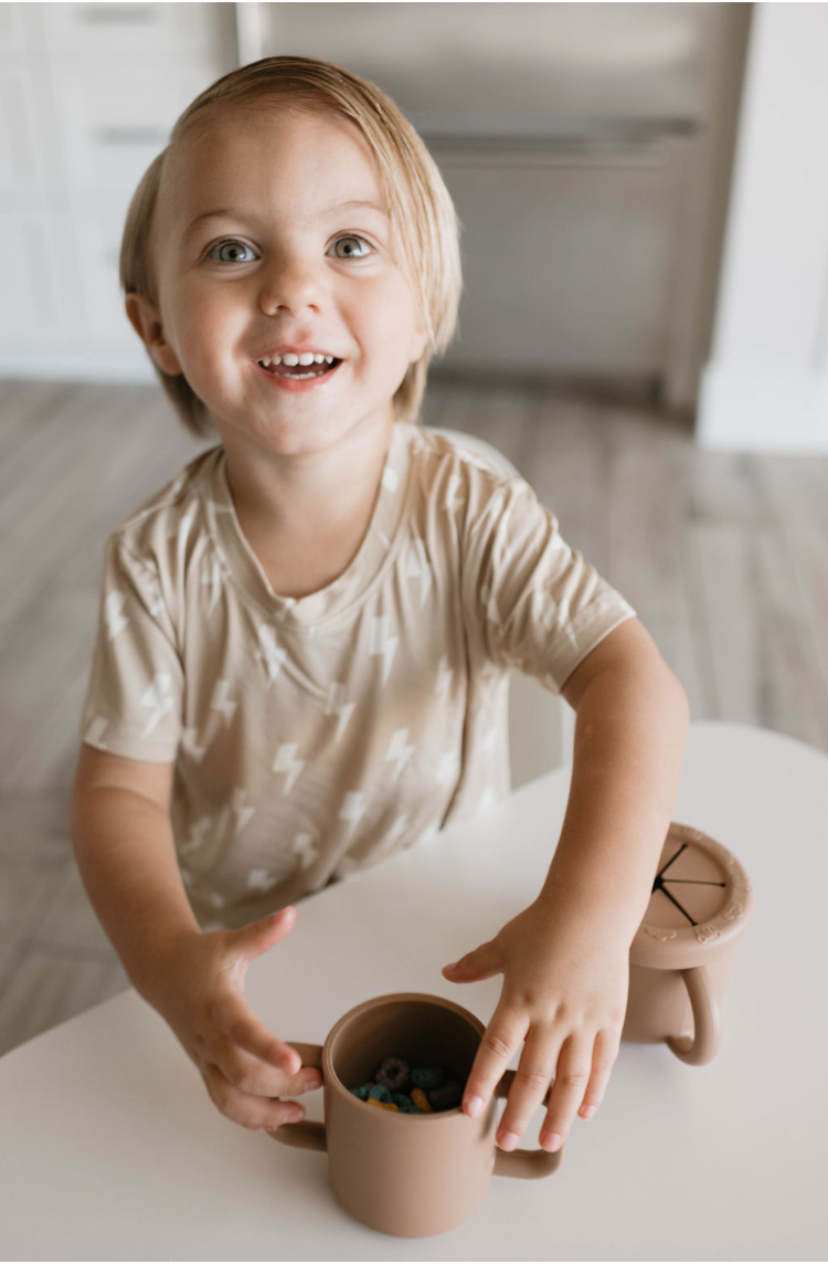 A young child with light brown hair is smiling and sitting at a table. The child is wearing a beige shirt with white lightning bolts and holds a BPA-free Forever French Baby Snack Cup with Removable Lid filled with fruit-flavored cereal. Another beige cup and a coaster are on the table, perfect for easy snacking.