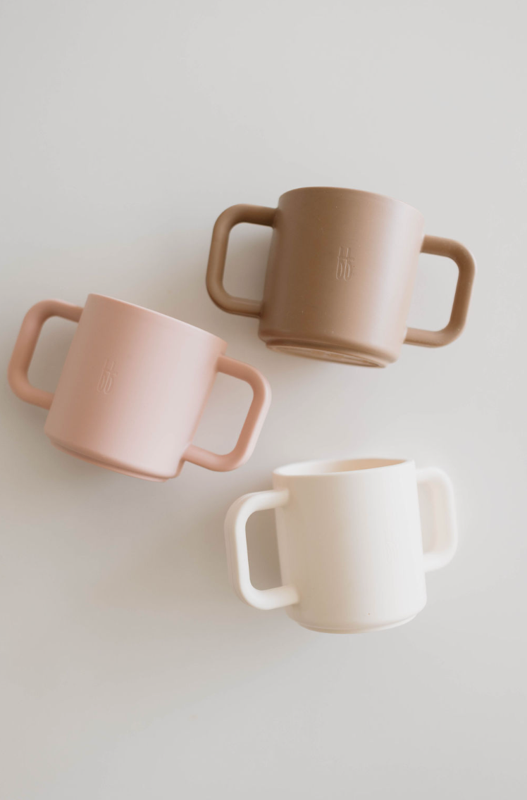 Three forever french baby Silicone Training Cups, crafted from BPA-free food safe silicone, are arranged on a white surface. The cups come in pastel shades of pink, brown, and cream. Each training cup features a double handle design for easy grip.