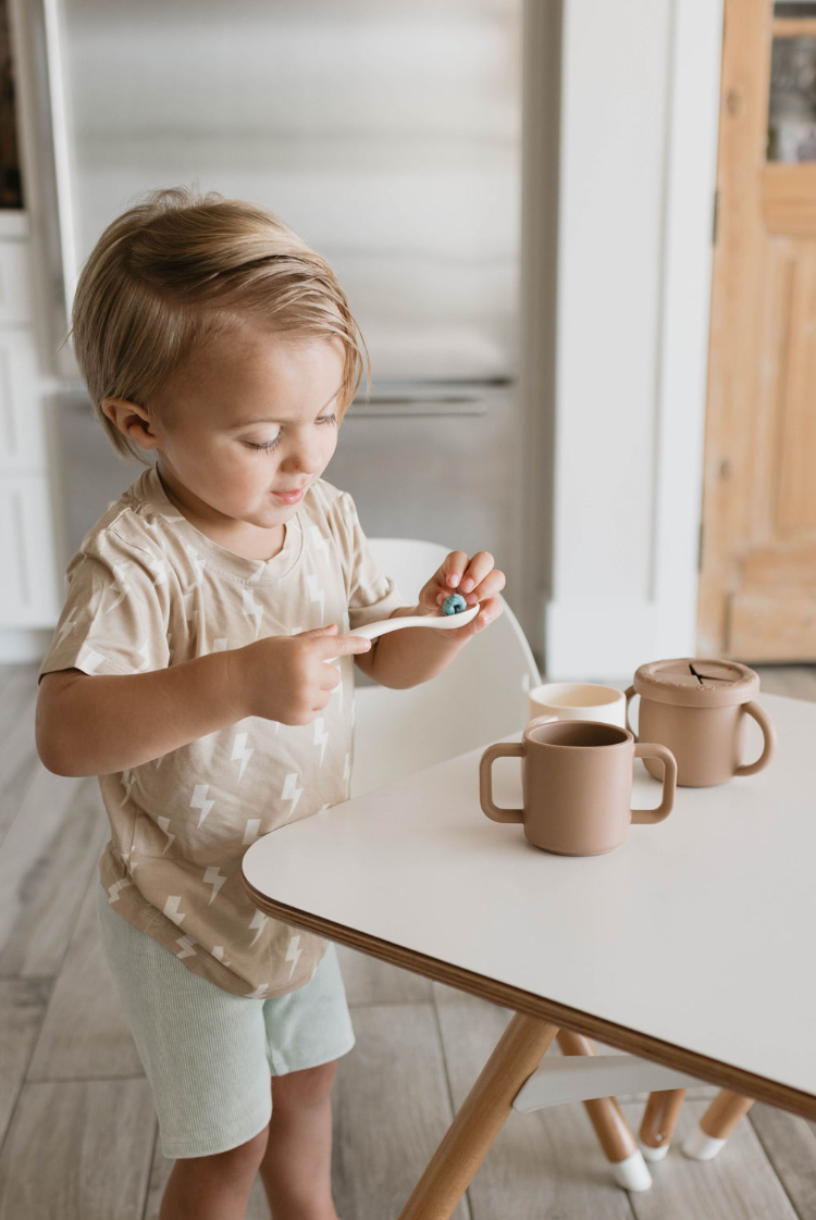 A young child with blond hair stands by a white table, using a spoon to stir a small item in a matching brown, BPA-free Silicone Training Cup from forever french baby. The child is dressed in a light brown shirt adorned with white lightning bolts and light-colored shorts. Two more cups are on the table, and there is a chair with wooden legs nearby.