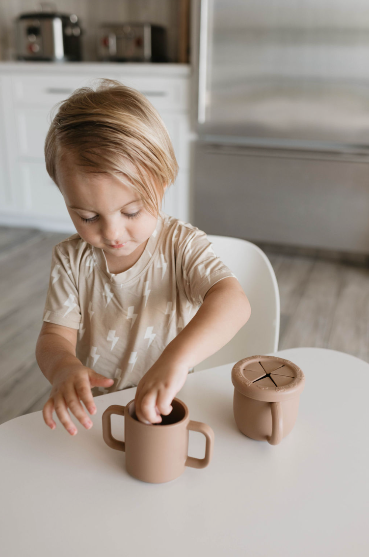 A young child with blond hair wearing a beige shirt is sitting at a white table in a kitchen. The child is playing with the forever french baby Silicone Training Cup, which is BPA-free and food-safe, and features handles for easy gripping. Next to the child on the table is another brown silicone cup from the same brand, this time with a lid.