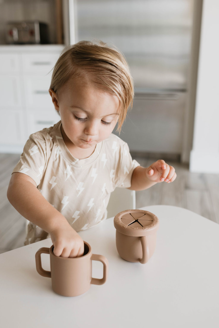 A small child with light brown hair is sitting at a white table in a modern kitchen. They are wearing a beige shirt with white lightning bolt patterns. The child is interacting with two BPA-free, food-safe silicone cups from forever french baby—one with a spout and another designed as the Silicone Training Cup featuring two handles.