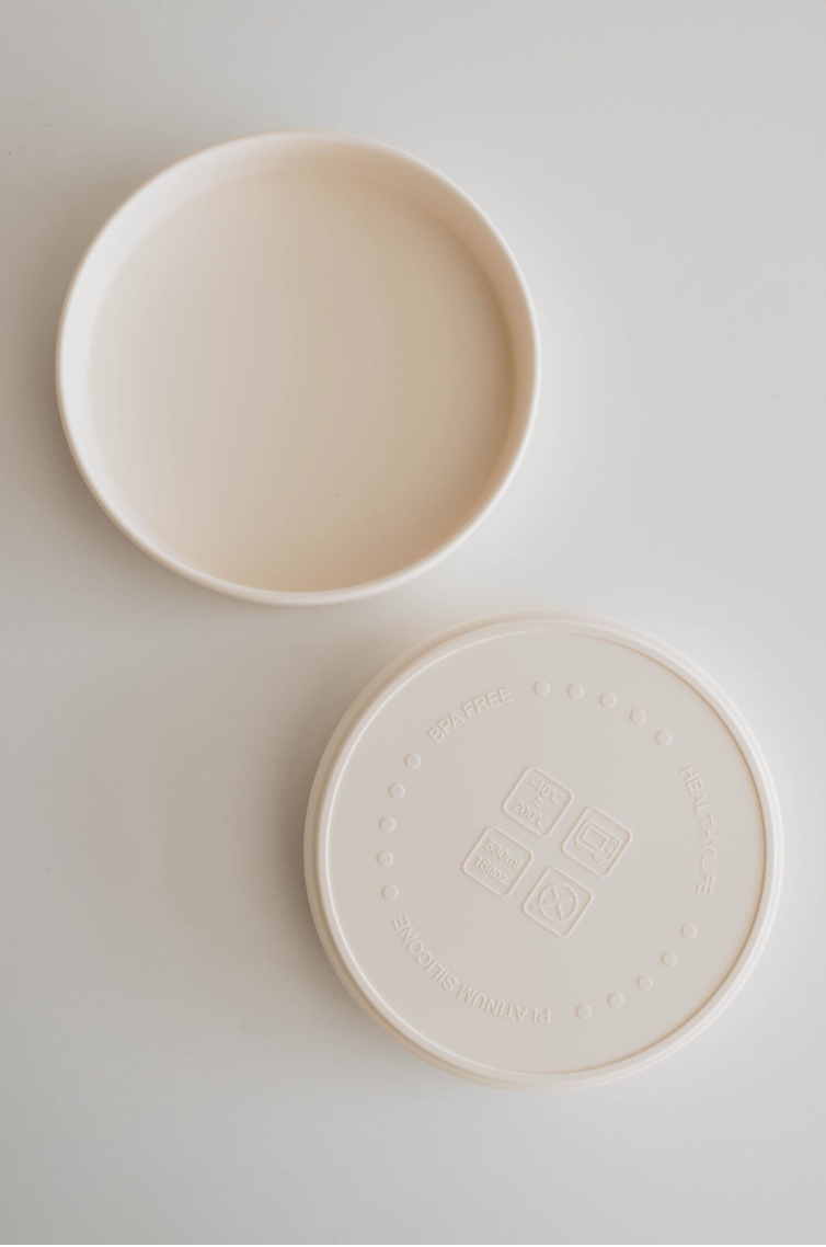 A round, ivory, BPA-free silicone Training Plate by forever french baby features both a lid and a base. The base is open-ended, while the lid is imprinted with symbols indicating it's safe for dishwashers, microwaves, and freezers—making it the perfect companion for mealtime.