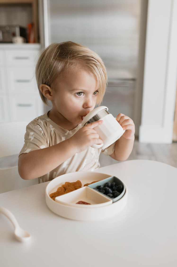 A young child with blonde hair sits at a white table, drinking from a white mug. In front of them is the food-safe silicone Training Plate | Ivory by forever french baby, divided into sections filled with blueberries, crackers, and possibly other snacks. The background showcases a modern kitchen setting, making mealtime both fun and convenient.