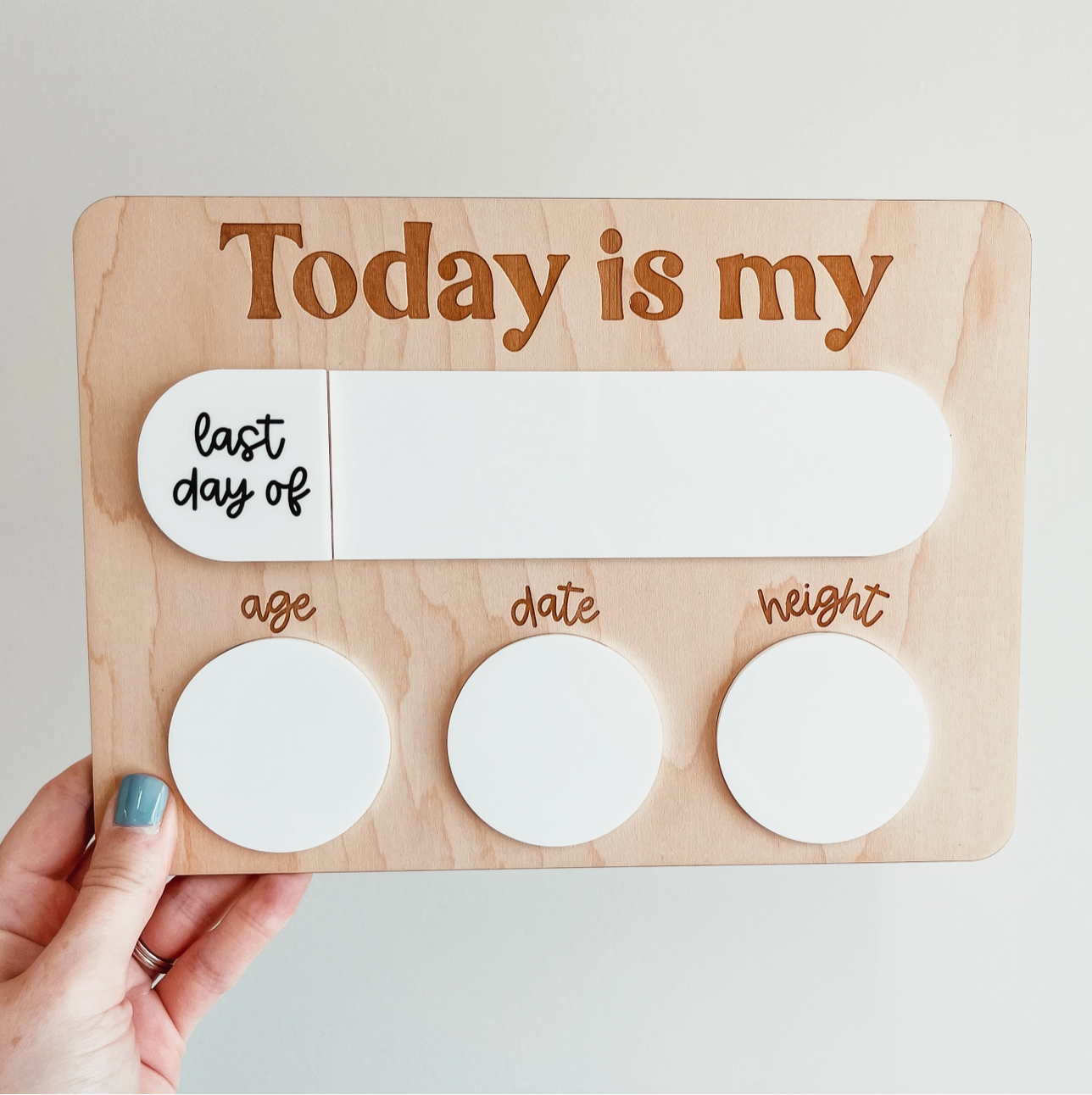 A hand holding the "First Day | Last Day School Sign" from Knotty Design Co, with the text "Today is my" engraved at the top. Below it, a white space labeled "last day of school" is perfect for back-to-school photos. Underneath, three circular white areas are labeled "age," "date," and "weight.