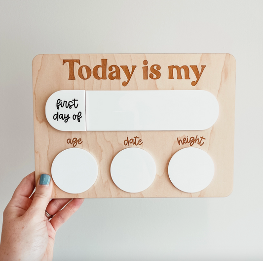 A hand with blue nail polish on the thumb is holding the Knotty Design Co's "First Day | Last Day School Sign." The wooden milestone board features the phrase "Today is my first day of" at the top, followed by three circular sections labeled "age," "date," and "weight," each with a white space to write information. This sign is perfect for back-to-school photos.