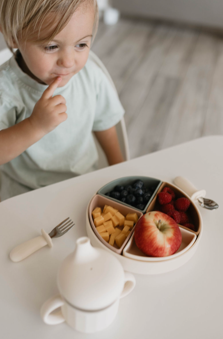 A young child sits at a table with a thoughtful expression, looking at a divided plate filled with blueberries, raspberries, cheese cubes, and an apple. In front of the plate is a sippy cup and a fork with a silicone handle from the forever french baby Training Utensil Set, perfect for self-feeding.