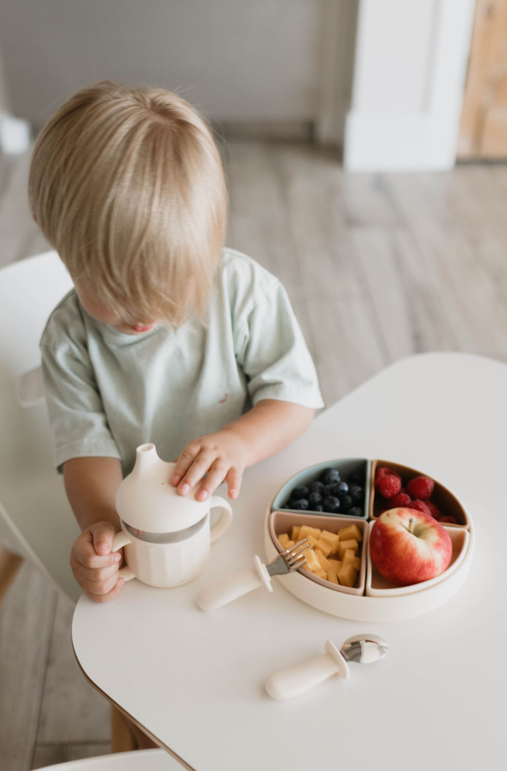 A toddler with blonde hair is seated at a white table, holding a sippy cup. In front of the child is a divided plate containing blueberries, diced cheese, raspberries, and an apple. The table also features a Training Utensil Set with silicone handles designed for self-feed training by forever french baby. The toddler is wearing a light-colored shirt.