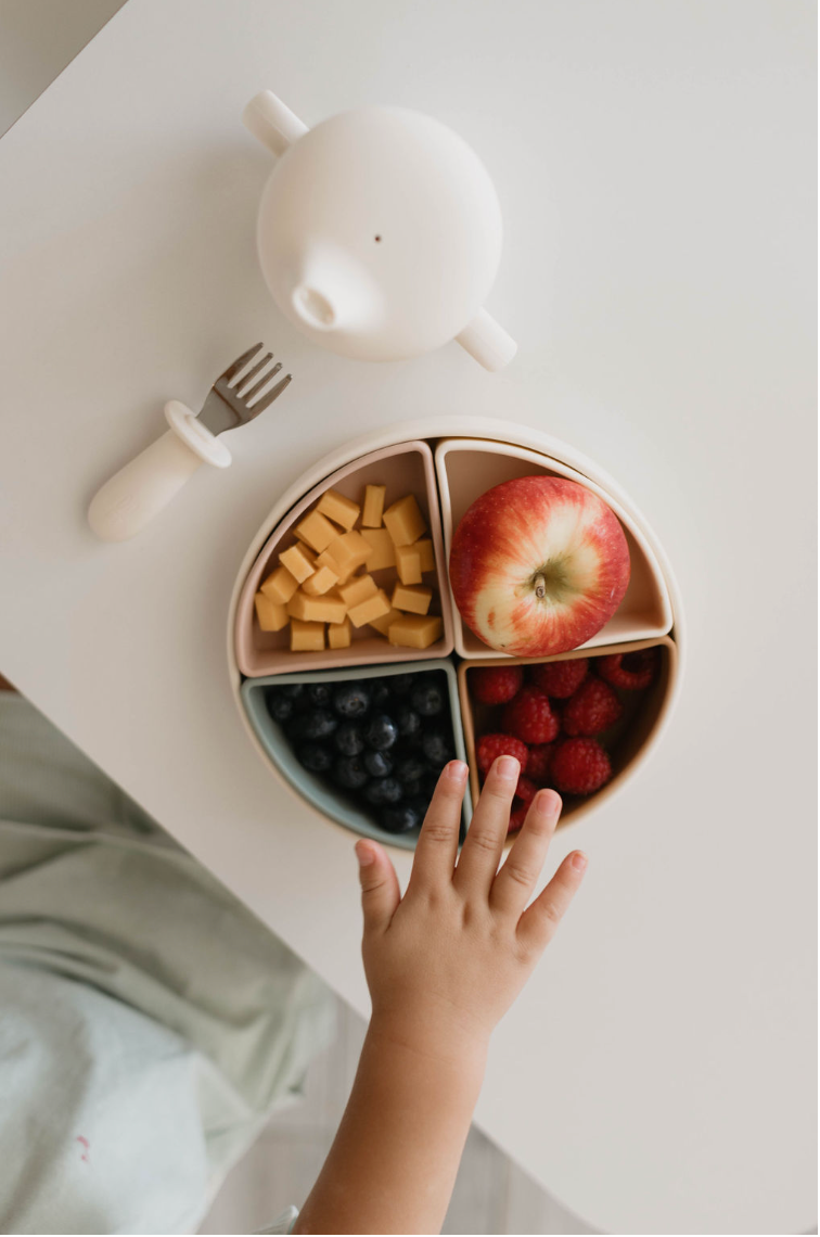 A child's hand reaches for a sectioned plate with an apple, cheese cubes, blueberries, and raspberries. Next to the plate on the white surface, there’s a small white plastic cup with a spout and a fork from our forever french baby Training Utensil Set featuring a silicone handle designed to encourage self-feeding.