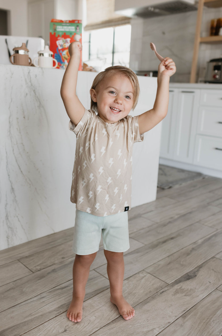 A cheerful, barefoot toddler with shoulder-length blonde hair stands in a bright, modern kitchen. The child is wearing a beige t-shirt with white lightning bolts and light-colored shorts, smiling widely with arms raised in excitement while holding the BPA-free Silicone Utensil Set from forever French baby.