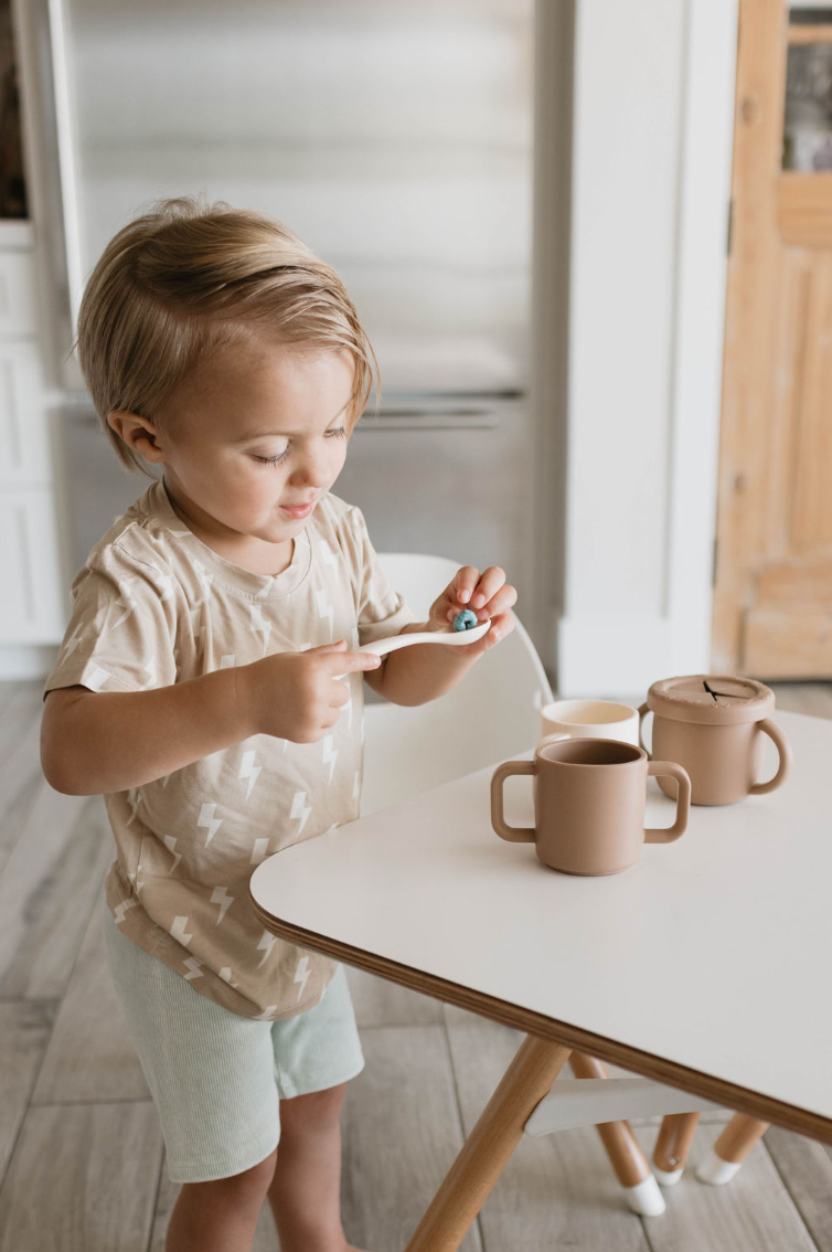 A young child stands at a white table holding a spoon with some blue food. The child is dressed in a beige shirt with white lightning bolt patterns and light-colored shorts. On the table, there are two brown toy cups, a toy teapot, and baby utensils made from BPA-free silicone from forever French baby's Silicone Utensil Set. The background features a kitchen scene.