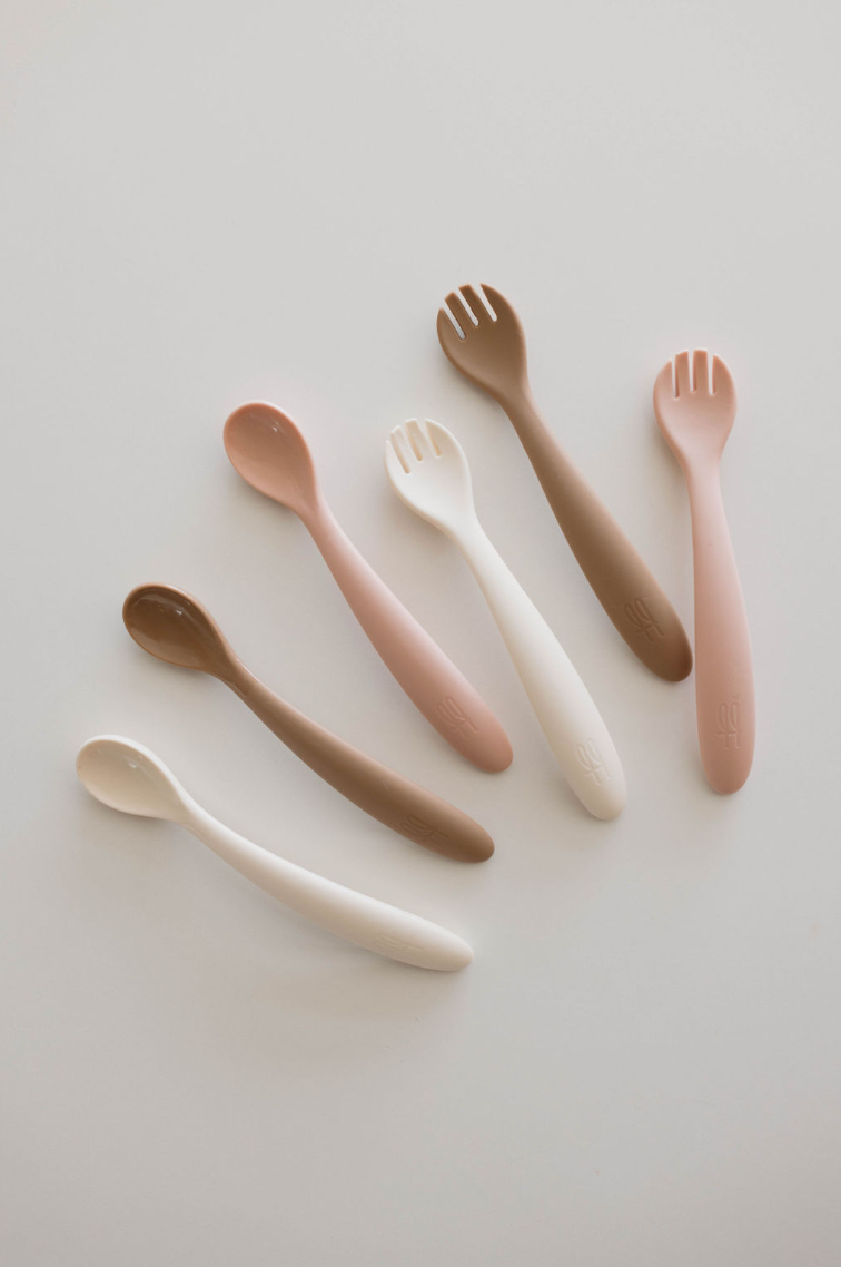 A set of seven BPA-free, silicone feeding utensils from forever French baby, including spoons and forks, laid out on a pale surface. The Silicone Utensil Set features soft shades of brown, pink, and white with smooth, rounded handles suitable for young children. The arrangement alternates between spoon and fork designs.