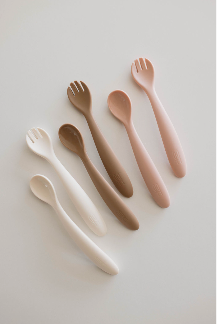 A set of six BPA-free feeding utensils from the Silicone Utensil Set by forever French baby, arranged in two rows against a white background. The baby utensils vary in colors ranging from cream, brown, beige, and off-white to light pink. Each spoon has a smooth handle, with the top-row spoons featuring spork-like prongs.