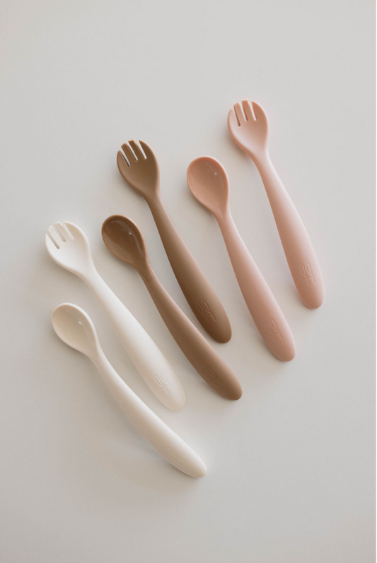 A set of six BPA-free feeding utensils from the Silicone Utensil Set by forever French baby, arranged in two rows against a white background. The baby utensils vary in colors ranging from cream, brown, beige, and off-white to light pink. Each spoon has a smooth handle, with the top-row spoons featuring spork-like prongs.