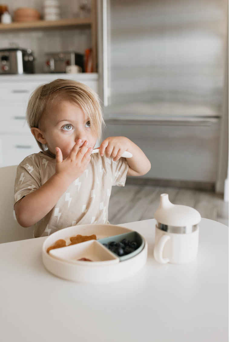 A young child with light hair sits at a table, wearing a tan shirt with a lightning bolt pattern. The child holds a spoon in their mouth, looking upwards. In front of them is a divided plate with snacks and the Silicone Utensil Set from forever French baby, along with a sippy cup. The background features a kitchen.