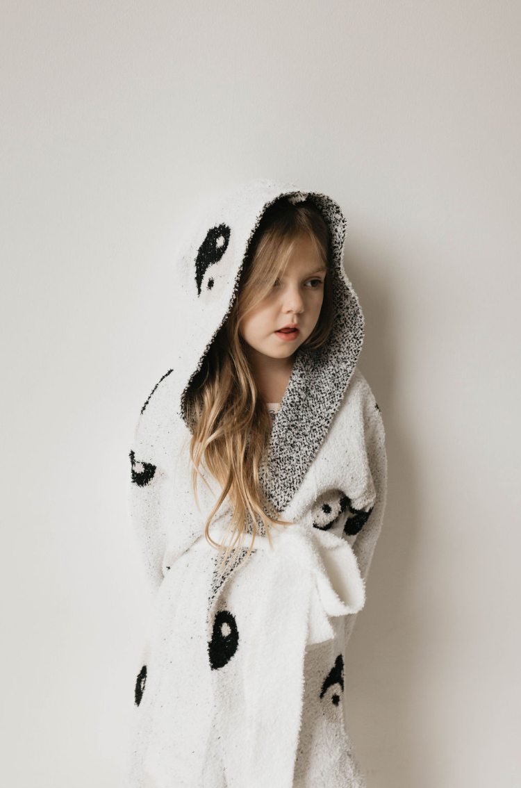 A young child with long, blonde hair wears a white hooded robe featuring yin-yang symbols from Forever French's Children's Robe | Yin Yang collection. Made from Oeko-tex certified materials, the robe exemplifies quality and comfort. The child stands against a plain, light-colored background, looking slightly to the side with a calm expression.