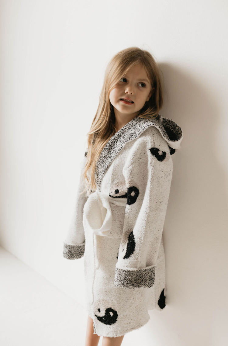 A young girl stands against a white wall, wearing the Children's Robe | Yin Yang from Forever French. The fluffy bathrobe, made of microfiber feather yarn with black and gray yin-yang symbols, features a hood and is belted at the waist. Her long, straight hair falls over her shoulders as she looks slightly to the side.