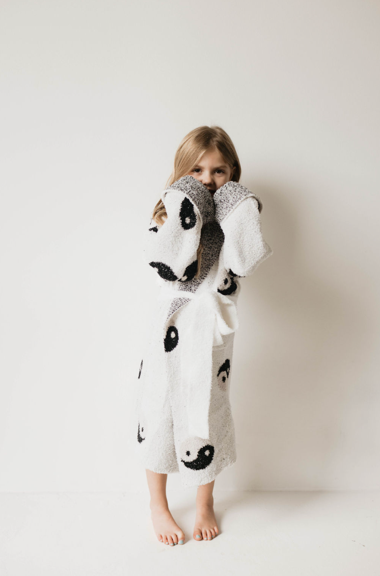 A young girl stands against a white background, wrapped in a fluffy white robe featuring a panda design. Crafted from Forever French's microfiber feather yarn and Oeko-tex certified for ultimate softness and safety, the **Children's Robe | Yin Yang** offers unparalleled comfort. She has long, light brown hair and is barefoot, looking shyly at the camera with her hands partially covering her face.