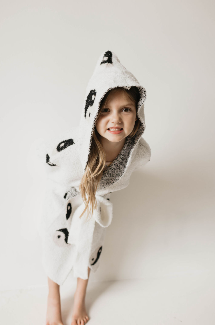 A young girl with long hair is wearing the Children's Robe | Yin Yang made from soft microfiber feather yarn with a playful panda design. She is standing barefoot and leaning slightly forward, looking at the camera with a small smile. The Oeko-Tex certified robe from Forever French highlights her cute expression and fun attire.