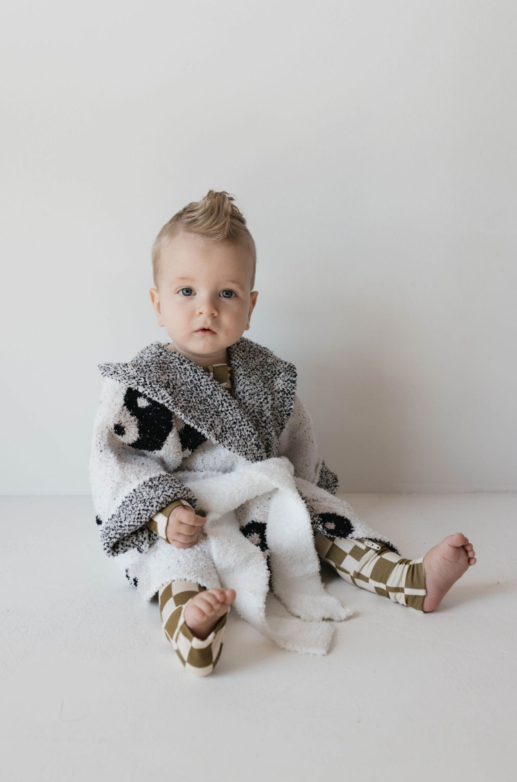 A baby sits against a white background, wearing a Forever French Baby patterned outfit and Children's Robe | Yin Yang, which features black spots on fluffy white fabric. Made of Oeko-tex certified materials, the baby's short blonde hair is styled in a small mohawk as they look directly at the camera with a calm expression.