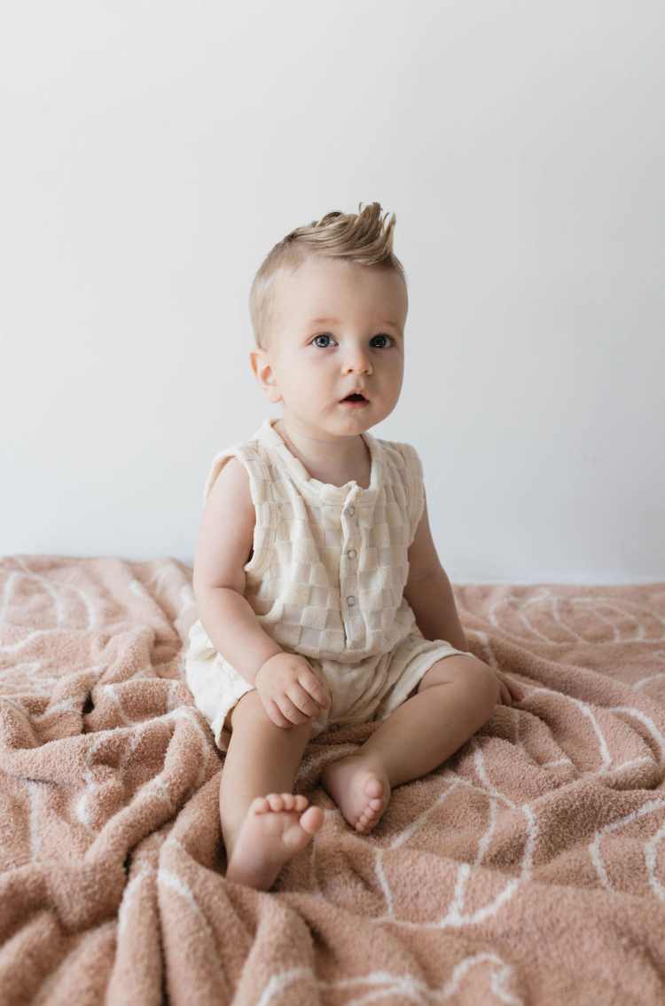 A baby with light hair sits on a soft, patterned blanket. The baby is wearing the forever french baby Tank Romper in Cream Checkerboard, which features snap closures down the front. The background is plain white.