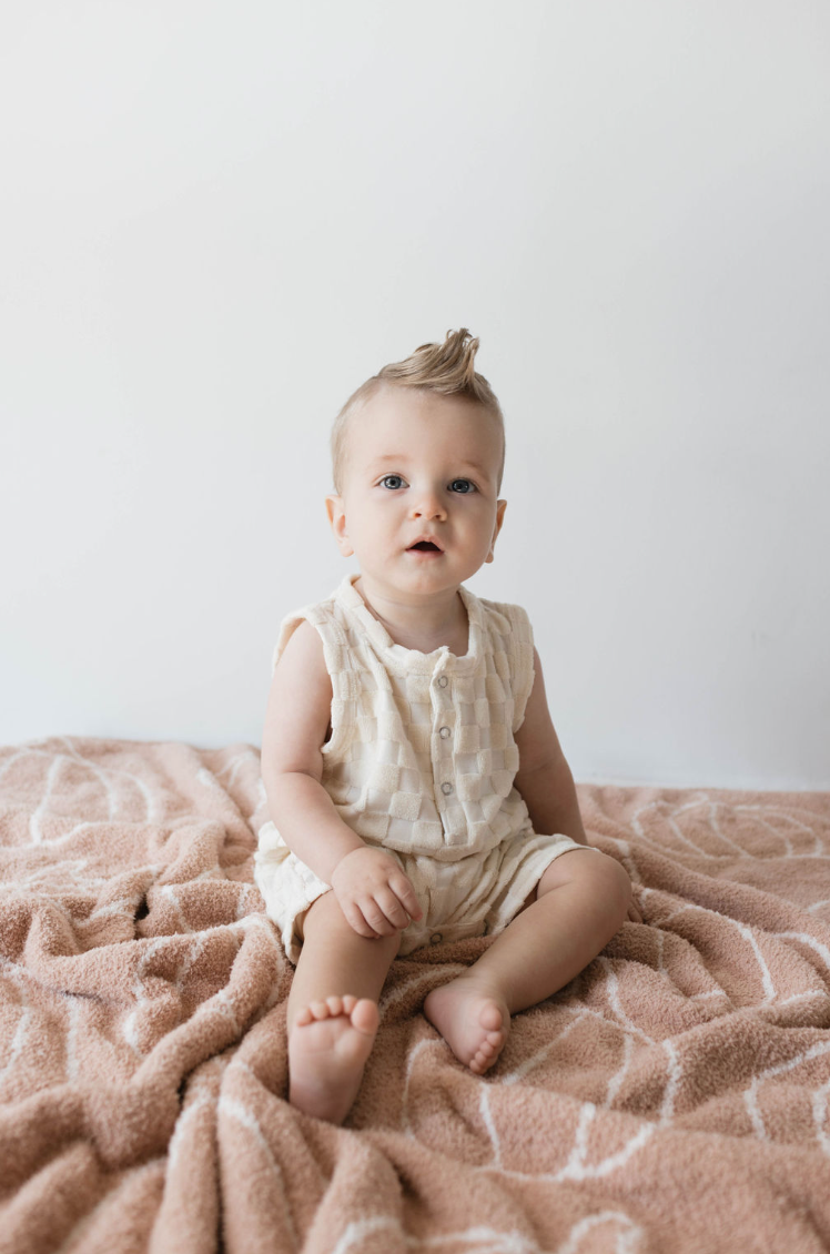 A baby with blonde hair sits on a peach-colored textured blanket. The little one is dressed in a Forever French Baby Tank Romper in Cream Checkerboard, featuring snap closures, and is looking forward with a neutral expression. The background is plain white.