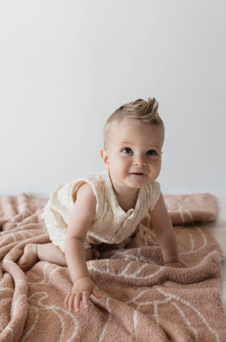 A baby with short, light hair and dressed in a sleeveless Tank Romper in Cream Checkerboard by forever french baby is crawling on an ultra-soft Terry patterned blanket. The baby has a curious expression and is positioned against a simple, light-colored background, reminiscent of a Forever French Baby setting.