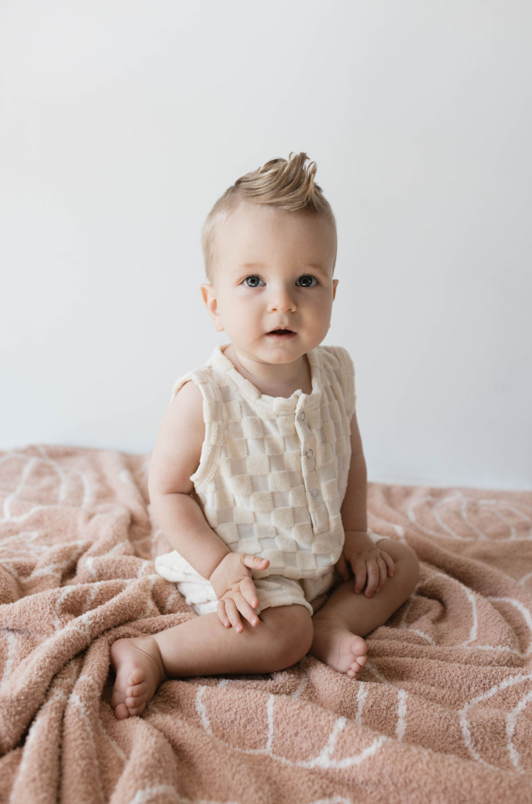 A baby with light hair sits on an ultra-soft pink textured blanket. The baby, dressed in a sleeveless Tank Romper | Cream Checkerboard from forever french baby, looks curious as snap closures ensure comfort. The plain white background draws attention to the baby's expression and relaxed posture.