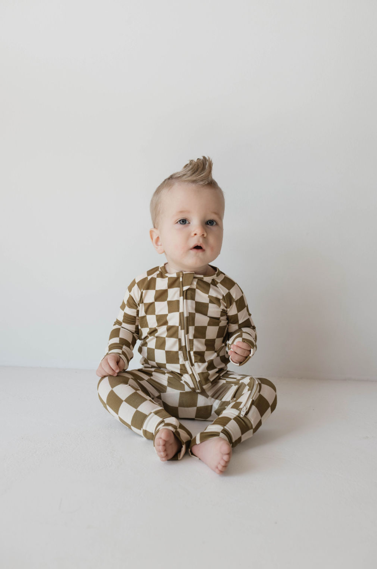 A baby with light-colored hair sits on the floor against a plain white background, dressed in premium Bamboo Zip Pajamas in an elegant Olive Checkerboard pattern from forever french baby. With a tuft of hair styled upwards, the baby gazes slightly to the right with a curious expression.