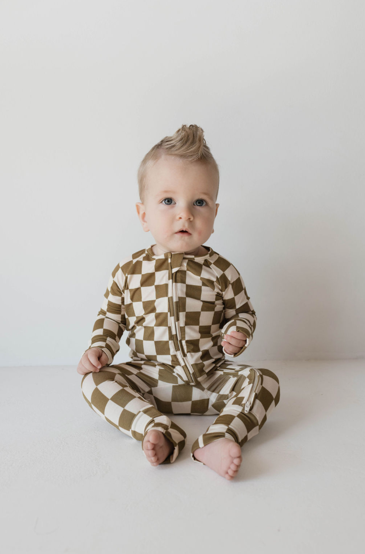 A baby with light-colored hair, styled in a small mohawk, sits against a white background. Wearing hypo-allergenic clothing that includes the Bamboo Zip Pajamas in Olive Checkerboard from forever french baby, the baby looks directly at the camera with a neutral expression.