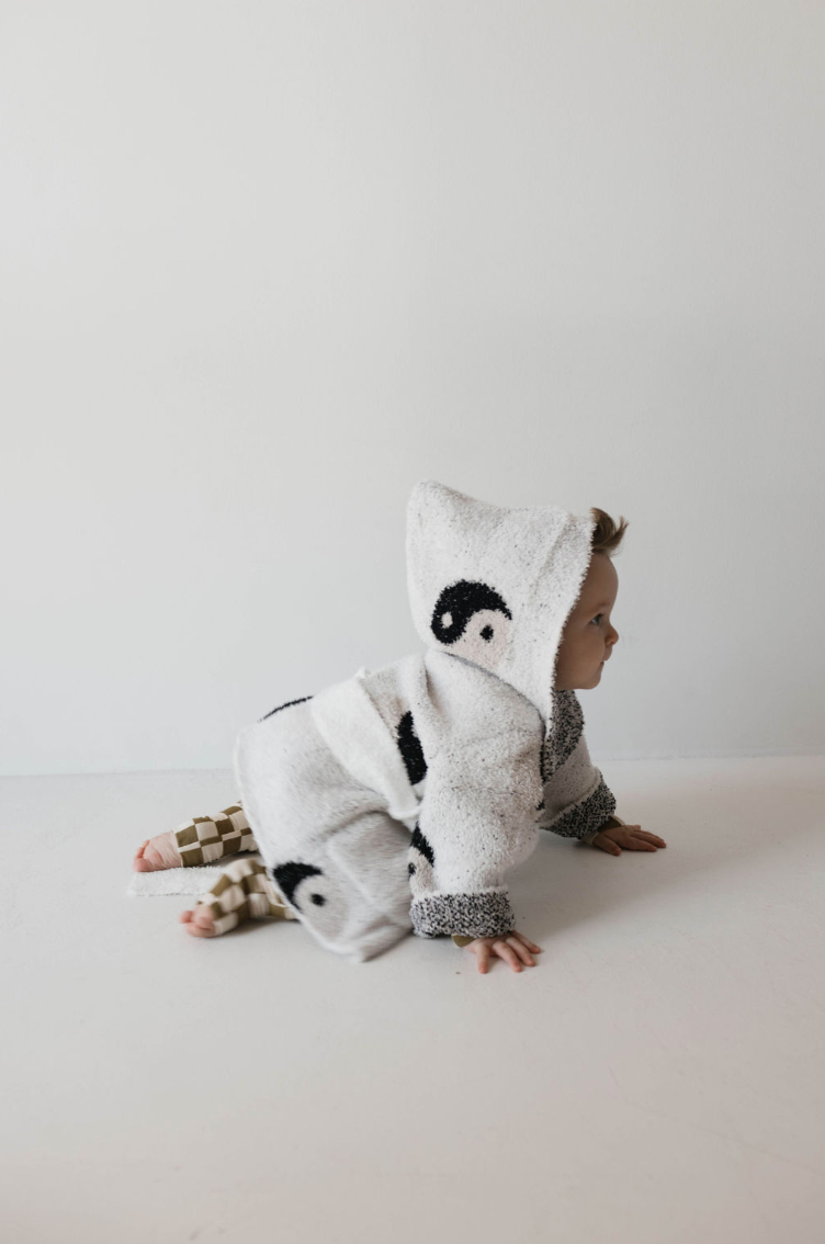 A baby dressed in a cozy, hooded, black and white Yin Yang-themed outfit from Forever French is crawling on a clean, white floor against a plain, light-colored wall. The OEKO-TEX certified outfit is made from Microfiber feather yarn, ensuring ultimate softness. The baby is looking off to the side.