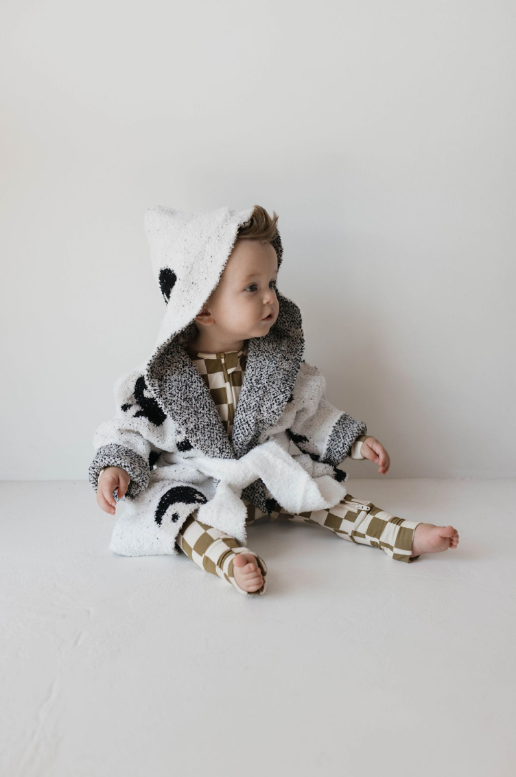 A young child sits on the floor wearing the Forever French Children's Robe in the Yin Yang design, featuring a cozy hood and made of microfiber feather yarn with a black and white spotted pattern. Underneath, the child sports a brown and white checkered outfit. The child is barefoot and gazes to the side with a curious expression.