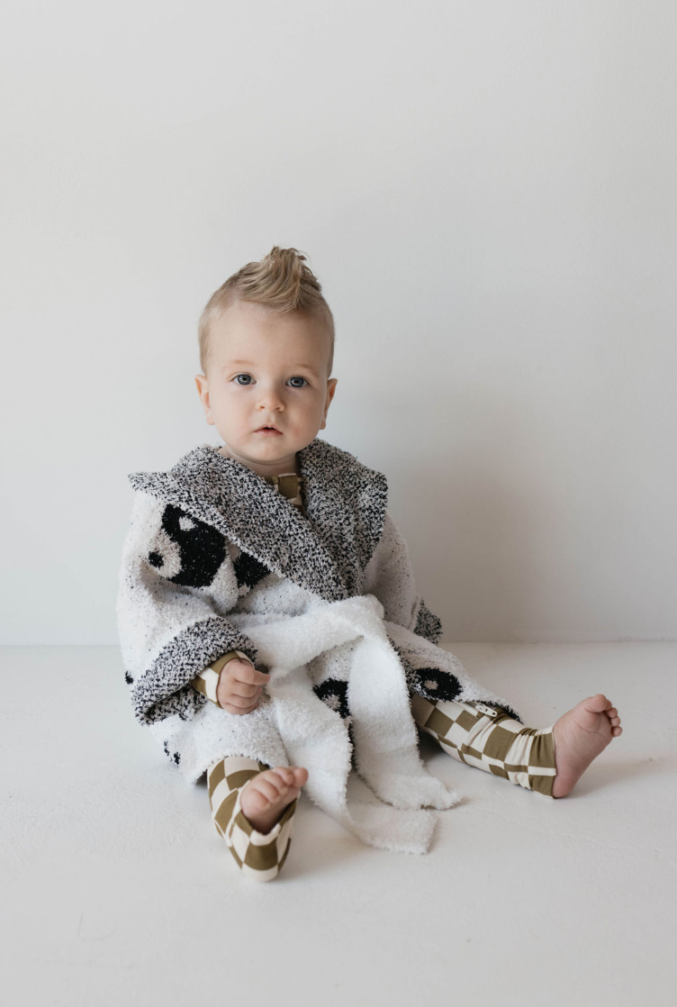 A baby with light hair sits against a plain white background, wearing a cozy Forever French Adult Robe | Yin Yang and patterned leggings made from soft Microfiber feather yarn. The baby looks directly at the camera with a calm expression.