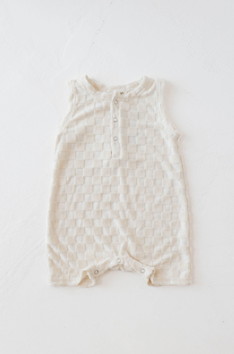 A Tank Romper in Cream Checkerboard from the forever french baby collection is laid flat on a light surface. This sleeveless romper features a small placket of buttons on the chest and snap closures at the bottom for easy diaper changes, with the fabric showcasing a soft, textured appearance.