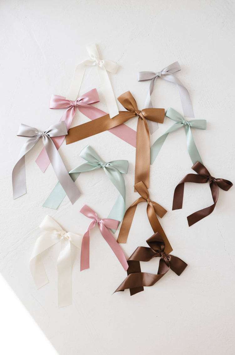 The "Hair Bow Clip | Satin" collection from forever french baby features ten carefully crafted satin bows in an assortment of colors, such as white, pink, brown, silver, mint green, and pastel blue. Each bow is meticulously designed and attached to an alligator clip. The bows are artistically scattered on a pristine white background.