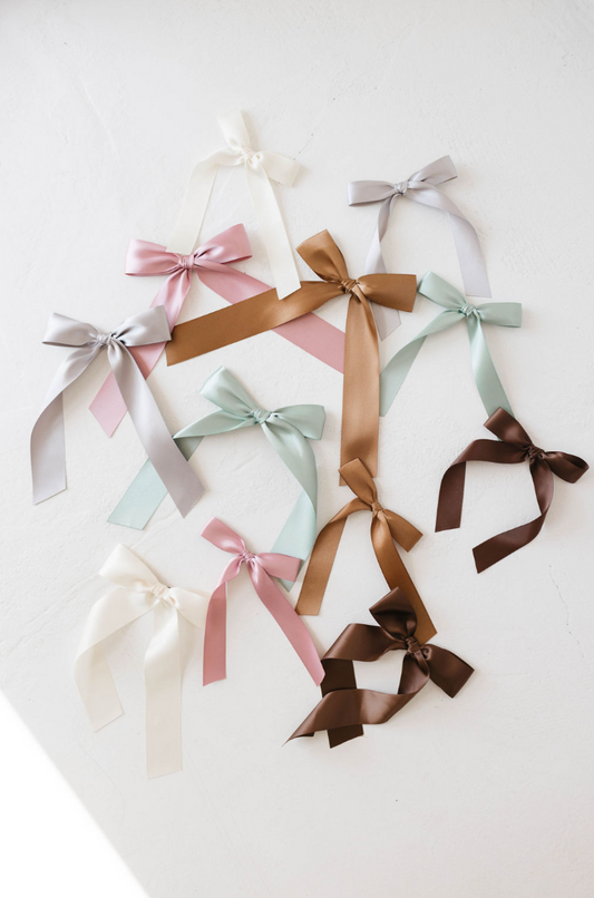 The "Hair Bow Clip | Satin" collection from forever french baby features ten carefully crafted satin bows in an assortment of colors, such as white, pink, brown, silver, mint green, and pastel blue. Each bow is meticulously designed and attached to an alligator clip. The bows are artistically scattered on a pristine white background.