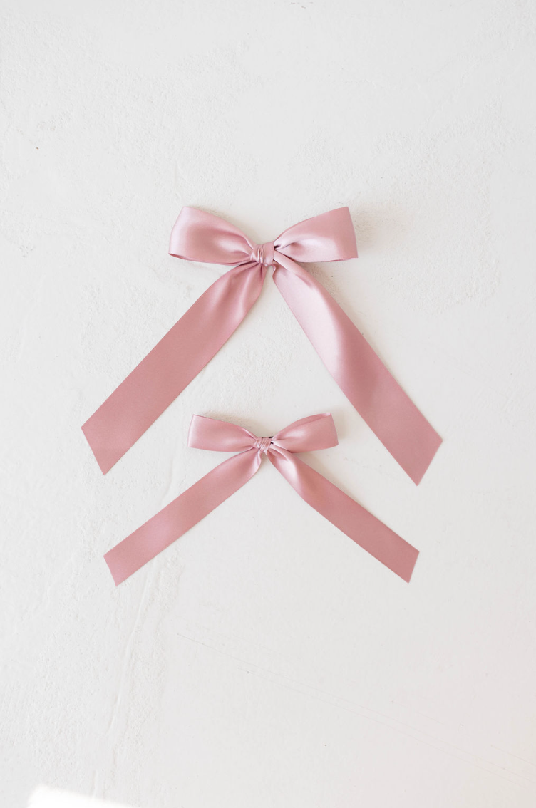 On a white surface, two satin hair clips from forever french baby are elegantly laid out. The top Hair Bow Clip | Satin showcases a larger bow with longer trailing ribbons, while the bottom one features a smaller bow with shorter ribbons. Both pink bows are neatly tied and exhibit a shiny, smooth appearance.