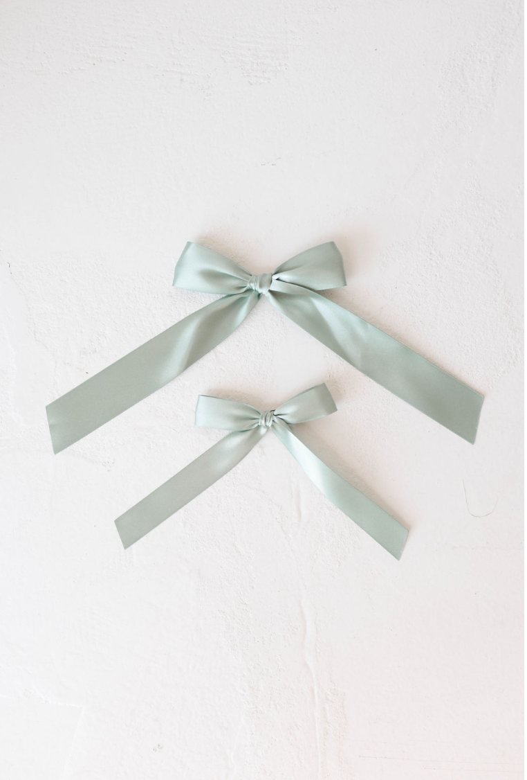 Two light green Hair Bow Clips by forever french baby, crafted from satin in different sizes, are laid flat on a white textured surface. The larger bow is positioned above the smaller one, both showcasing their tied ribbon loops and trailing ends, perfect for an elegant accessory.