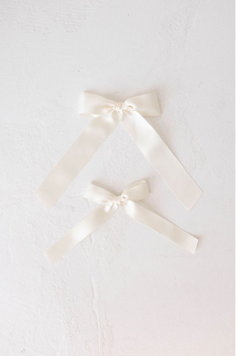 Two satin "Hair Bow Clips" in cream from forever french baby are arranged on a white, textured surface. Both clips feature smoothly tied loops and long, tapered tails, reminiscent of a classic bow design. The larger clip is placed above and slightly to the left of the smaller one. The scene is highlighted with soft lighting and maintains a minimalist aesthetic.