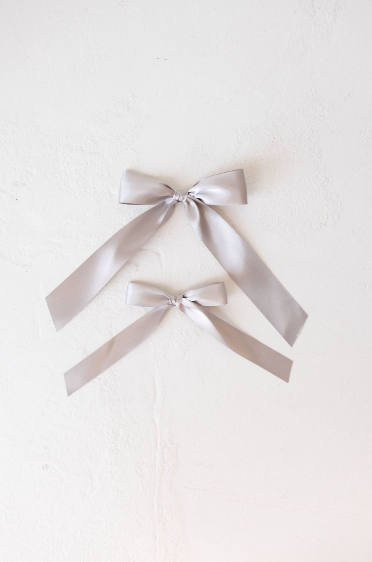 Two hair bow clips from forever french baby, made of luxurious silver satin, are laid flat on a white textured surface. The bows, one slightly larger than the other, each have long tails that drape down. This elegant design perfectly showcases the smooth and shiny texture of the satin fabric, making it ideal for satin hair clips or alligator clips.