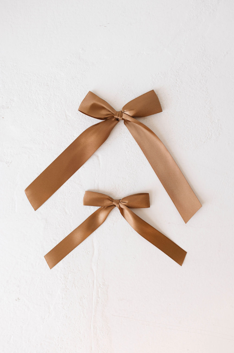 Two tied Hair Bow Clips in brown satin by forever french baby, perfect for a satin hair clip, are placed against a light textured background. The larger clip is above the smaller one, both forming neat bow designs with long tails hanging down.