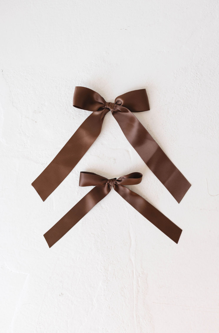 Two Hair Bow Clips | Satin by forever french baby, each in a beautiful brown satin fabric, are set against a white textured background. The clips are aligned vertically with the larger bow on top and the smaller one beneath. Both bows feature long tails and are meticulously tied, highlighting their smooth texture and elegant design.