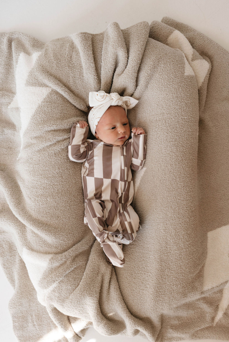 A newborn baby lies on a soft, beige blanket. The baby is wearing forever french baby's Bamboo Zip Pajamas in Tile Stack with a matching headband that has a bow on top. The baby's arms are slightly raised, resting against the cozy, plush layers of the blanket.