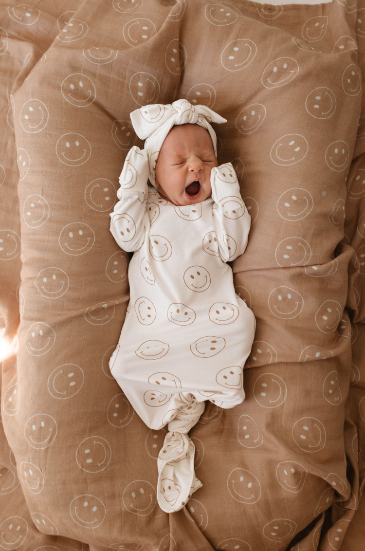 A newborn yawns on a brown blanket with white smiley faces, dressed in the forever french baby Bamboo Knotted Gown | Just Smile—an essential for gentle, comfortable care.