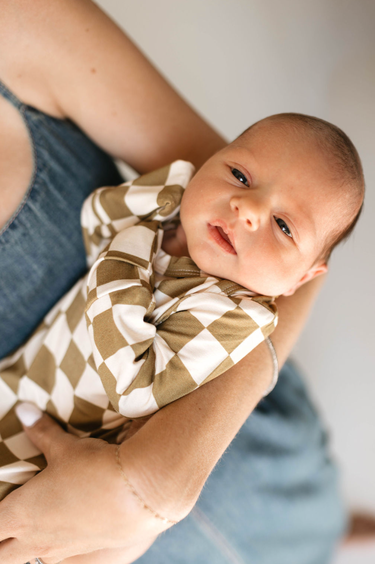 A newborn baby, dressed in a hypo-allergenic Olive Checkerboard Bamboo Knotted Gown by Forever French Baby, is being held in someone's arms. The baby's eyes are open and they appear calm and content. The person holding the baby is wearing a light-colored top and blue jeans.