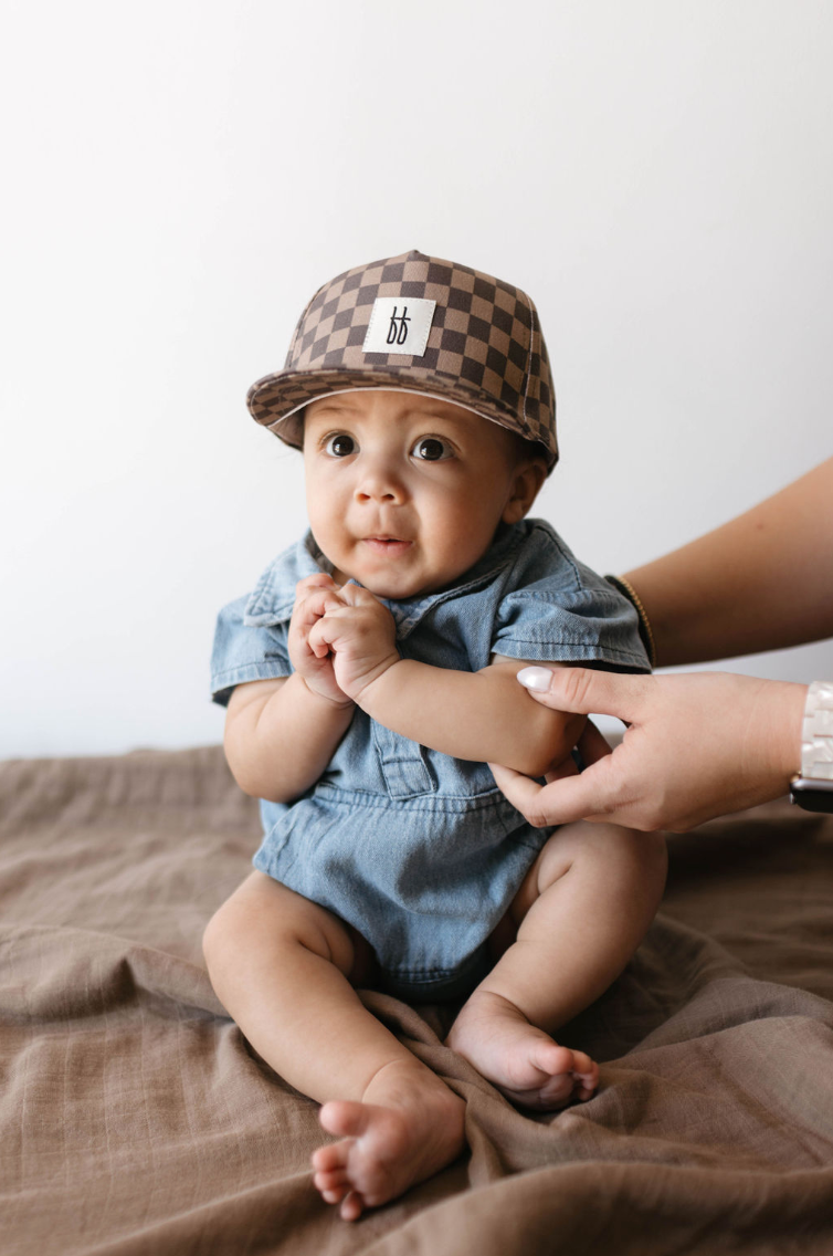 A baby wearing the Forever French Baby's Children's Trucker Hat in Faded Brown Checkerboard and a blue denim outfit sits on a brown blanket. An adult's hands gently support the baby from the side. The baby, with curious eyes and clasped hands, looks slightly upward, enjoying sun protection from their stylish cap.