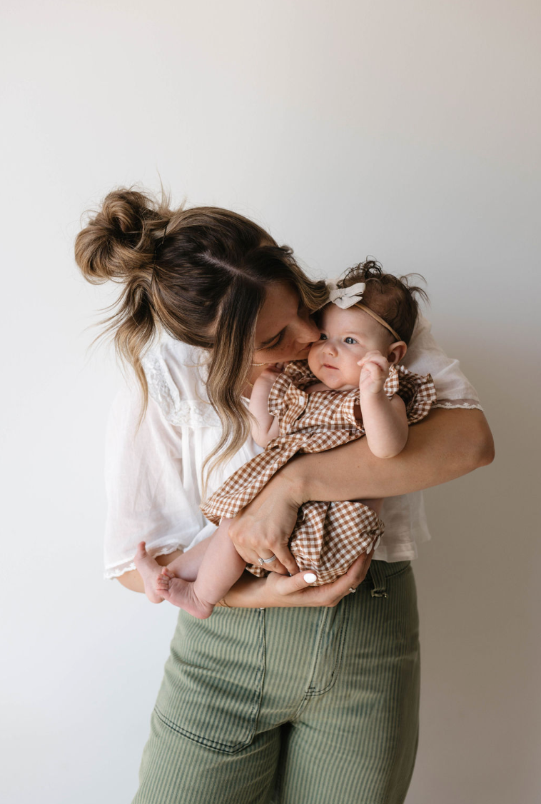 A woman with her hair in a bun, wearing a white blouse and green pants, lovingly embraces and kisses a baby. The baby, dressed in the adorable Gingham Bloomer Set | Honey from forever french baby with a small bow on their head, looks off into the distance. They stand against a plain white background.