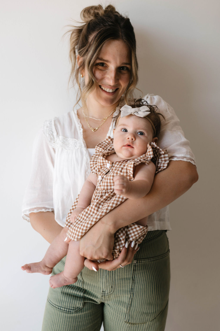 A woman with light brown hair tied back, dressed in a white blouse, cradles a baby adorned in the Gingham Bloomer Set | Honey from forever french baby. Both are smiling warmly against a simple grayish backdrop.