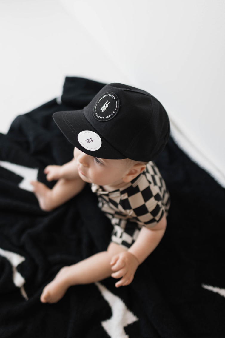 A baby sits on a black blanket, wearing the Children's Trucker Hat in ff Black by Forever French Baby and a black-and-white checkered outfit. The baby gazes up and to the side, softly lit by natural light in the background. Care instructions specify spot cleaning only.