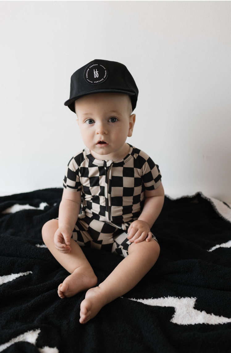 A baby wearing a Children's Trucker Hat | ff Black from forever french baby sits on a black and white patterned blanket against a plain white background, looking directly at the camera.