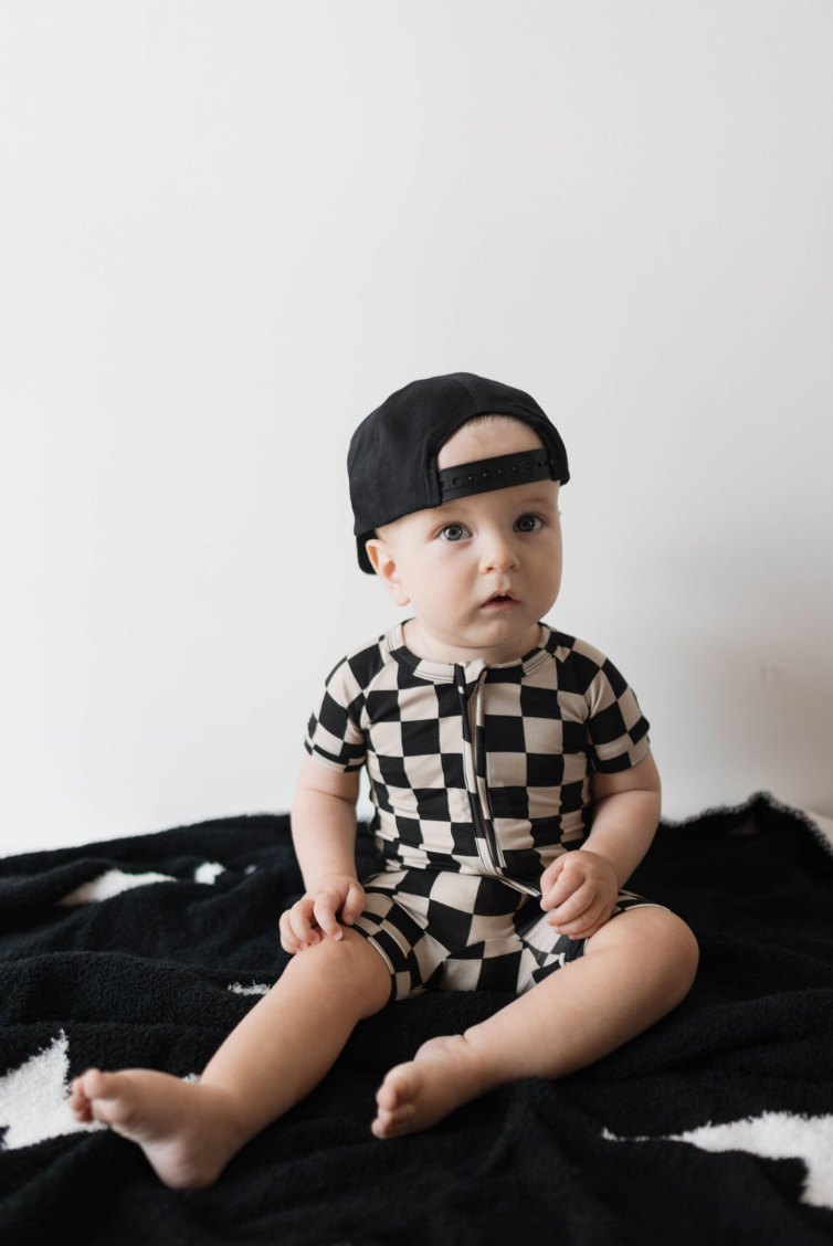 A baby sits on a black blanket against a plain background, wearing a black and white checkerboard outfit from Forever French Baby and a forever french baby ff Black Children's Trucker Hat worn backwards. The baby gazes forward with a curious expression, embodying timeless charm.